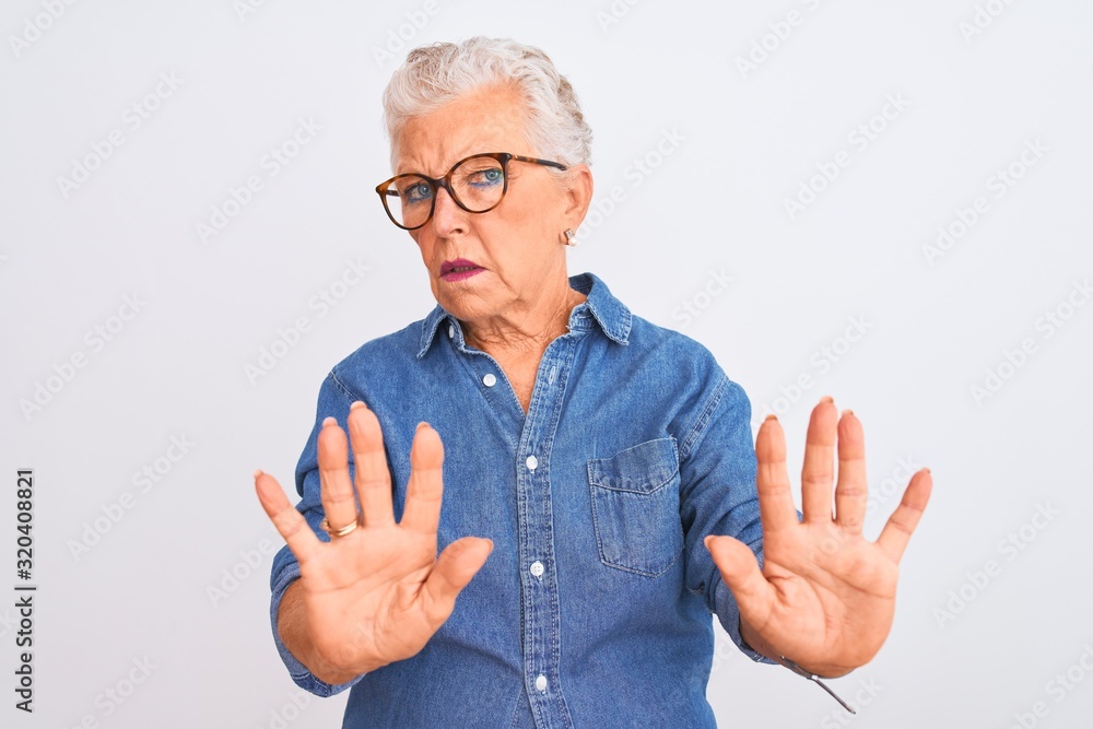 Senior grey-haired woman wearing denim shirt and glasses over isolated white background Moving away hands palms showing refusal and denial with afraid and disgusting expression. Stop and forbidden. Senior grey-haired woman wearing denim shirt and glasses over isolated white background Moving away hands palms showing refusal and denial with afraid and disgusting expression. Stop and forbidden.