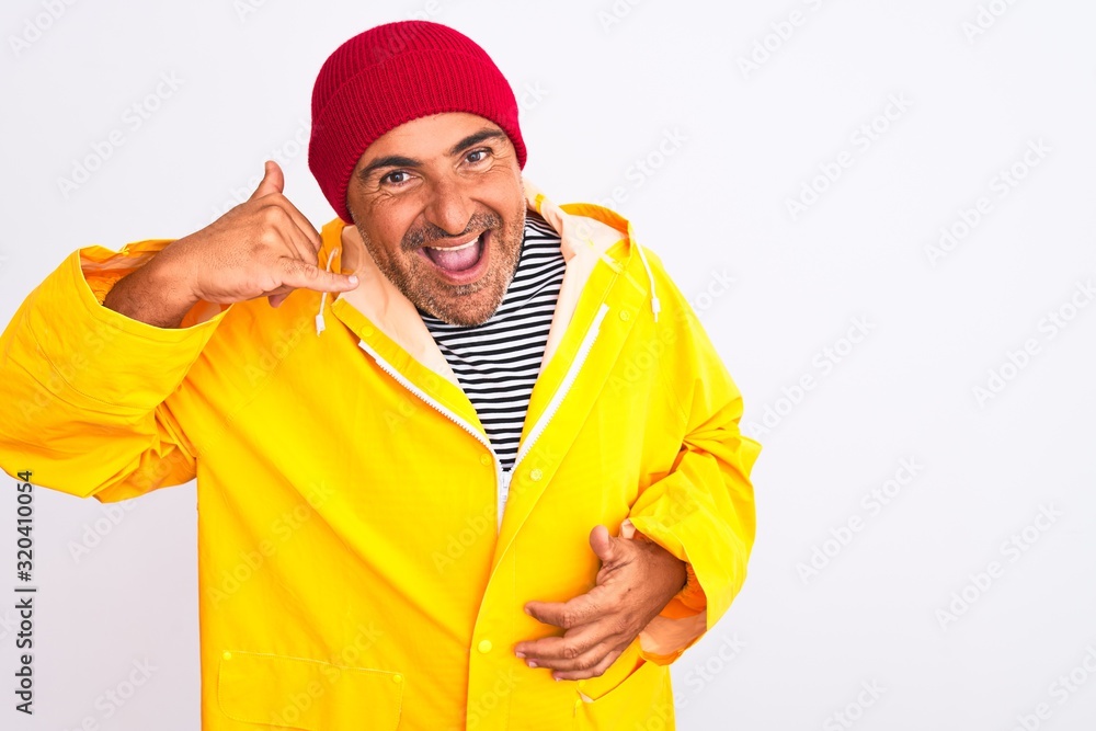 Middle age man wearing rain coat and woolen hat standing over isolated white background smiling doing phone gesture with hand and fingers like talking on the telephone. Communicating concepts.