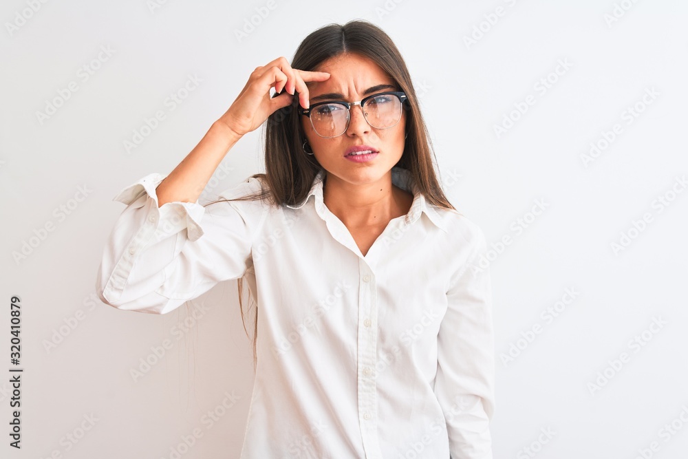 Young beautiful businesswoman wearing glasses standing over isolated white background pointing unhappy to pimple on forehead, ugly infection of blackhead. Acne and skin problem