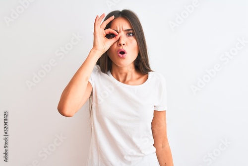 Young beautiful woman wearing casual t-shirt standing over isolated white background doing ok gesture shocked with surprised face, eye looking through fingers. Unbelieving expression.