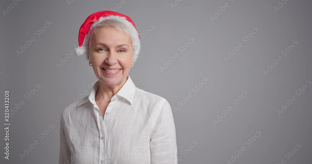 Attractive elderly woman in Santa hat smiling on gray background. Attractive elderly woman in Santa hat smiling on gray background.
