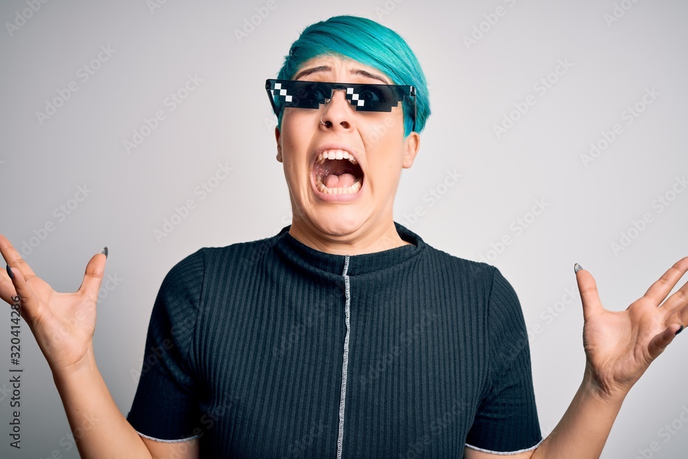 Young woman with blue fashion hair wearing thug life sunglasses over white background crazy and mad shouting and yelling with aggressive expression and arms raised. Frustration concept. Young woman with blue fashion hair wearing thug life sunglasses over white background crazy and mad shouting and yelling with aggressive expression and arms raised. Frustration concept.