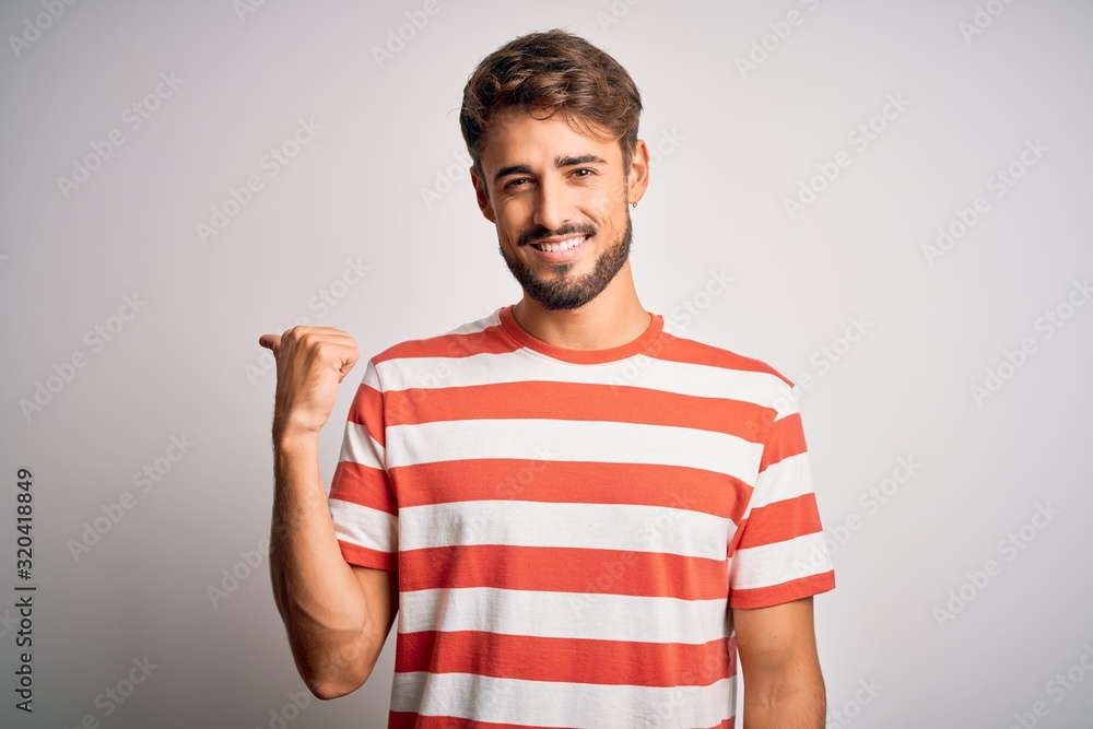 Young handsome man with beard wearing striped t-shirt standing over white background smiling with happy face looking and pointing to the side with thumb up. Young handsome man with beard wearing striped t-shirt standing over white background smiling with happy face looking and pointing to the side with thumb up.