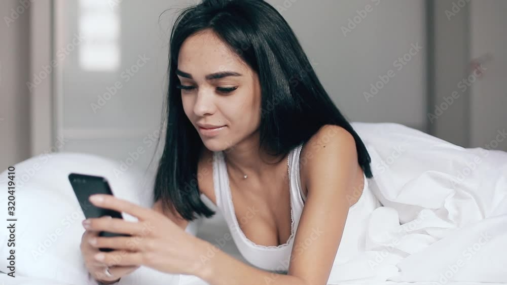 Pretty girl in bed reads messages on smartphone and smiles Pretty girl in bed reads messages on smartphone and smiles