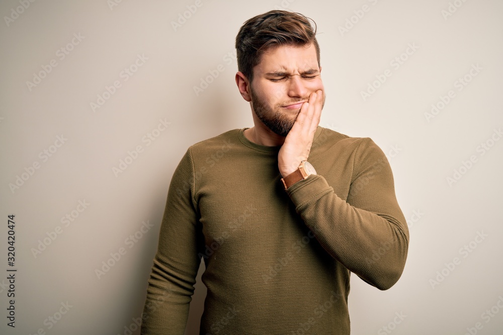 Young blond man with beard and blue eyes wearing green sweater over white background touching mouth with hand with painful expression because of toothache or dental illness on teeth. Dentist