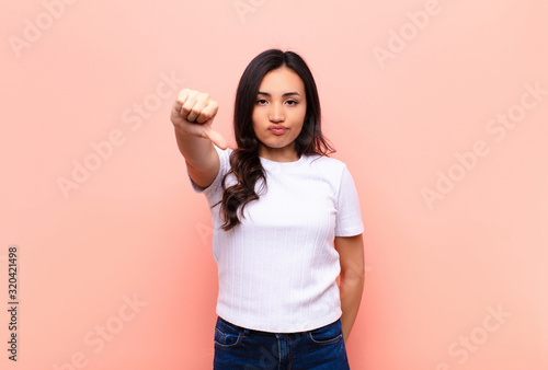 young latin pretty woman feeling cross, angry, annoyed, disappointed or displeased, showing thumbs down with a serious look against flat wall