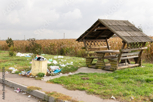 Full bin of rubbish with lots of waste around it by the wooden picnic table by the main motorway in Ukraine with cornfiled in the background. Waste in highways in eastern europe. photo