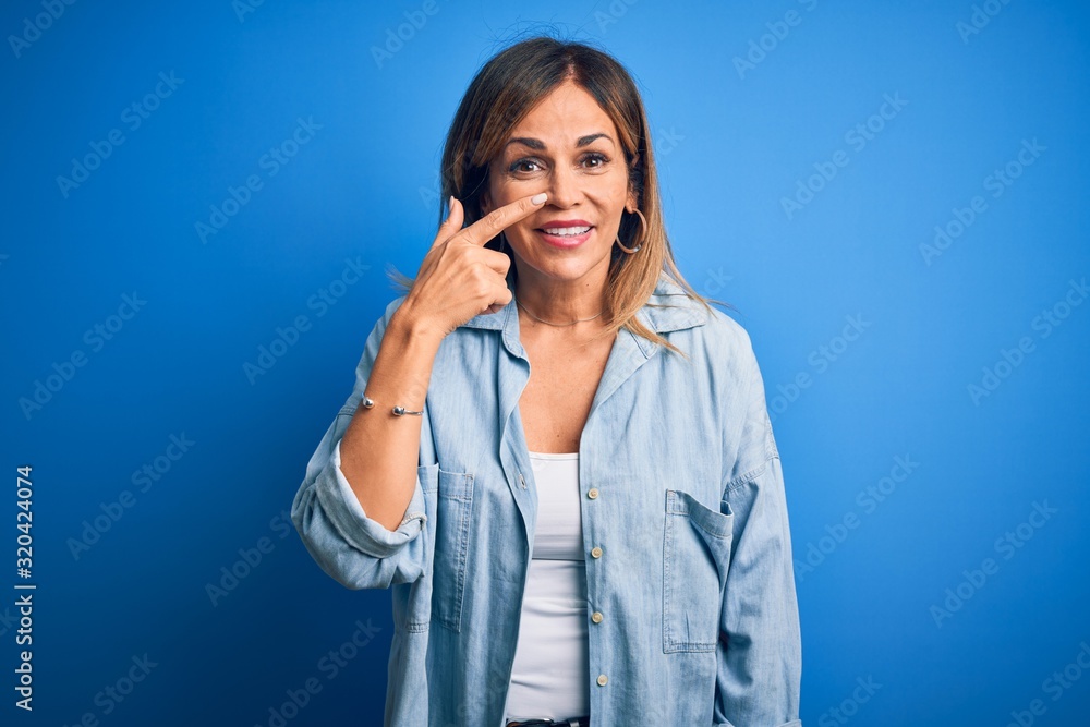 Middle age beautiful woman wearing casual shirt standing over isolated blue background Pointing with hand finger to face and nose, smiling cheerful. Beauty concept Middle age beautiful woman wearing casual shirt standing over isolated blue background Pointing with hand finger to face and nose, smiling cheerful. Beauty concept
