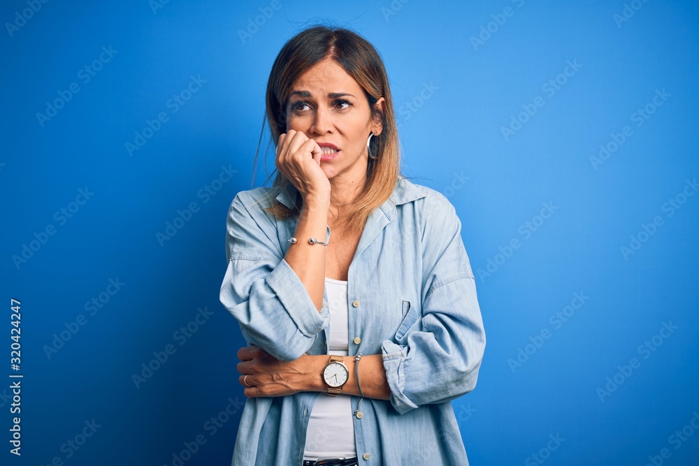 Middle age beautiful woman wearing casual shirt standing over isolated blue background looking stressed and nervous with hands on mouth biting nails. Anxiety problem. Middle age beautiful woman wearing casual shirt standing over isolated blue background looking stressed and nervous with hands on mouth biting nails. Anxiety problem.