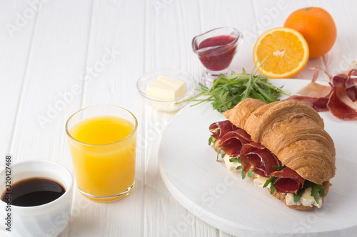 Fresh croissant sandwich with ham, cheese and arugula on white wooden table. Served with Coffee, cheese, ham, orange juice, jam. Fresh French Baked Croissants. Free space for text. 