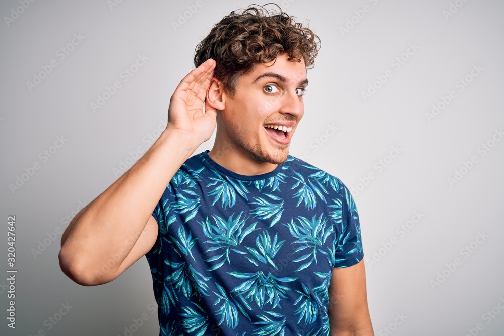Young blond handsome man on vacation with curly hair wearing casual summer t-shirt smiling with hand over ear listening an hearing to rumor or gossip. Deafness concept. Young blond handsome man on vacation with curly hair wearing casual summer t-shirt smiling with hand over ear listening an hearing to rumor or gossip. Deafness concept.