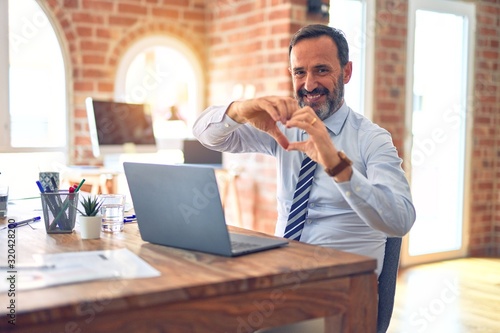 Middle age handsome businessman wearing tie sitting using laptop at the office smiling in love showing heart symbol and shape with hands. Romantic concept.