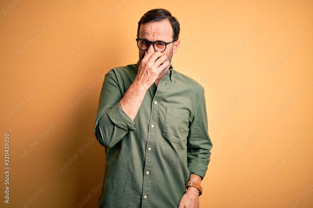 Middle age hoary man wearing casual green shirt and glasses over isolated yellow background smelling something stinky and disgusting, intolerable smell, holding breath with fingers on nose. Bad smell Middle age hoary man wearing casual green shirt and glasses over isolated yellow background smelling something stinky and disgusting, intolerable smell, holding breath with fingers on nose. Bad smell
