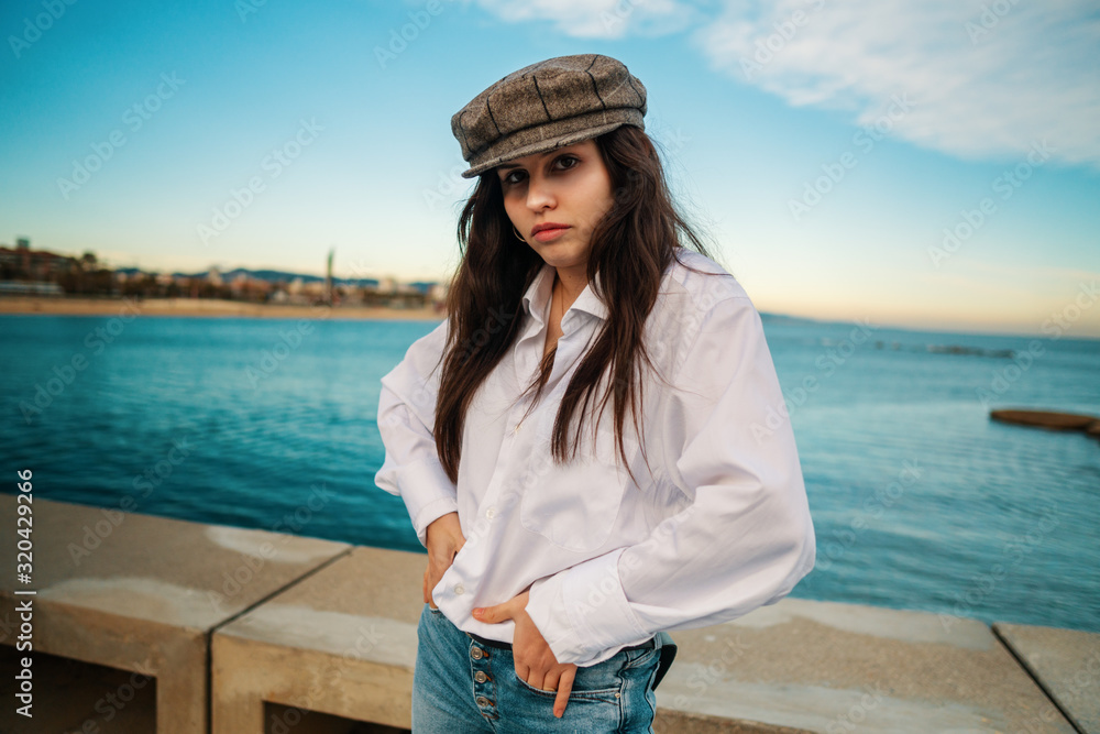 Portrait of young beautiful stylish woman in white shirt and grey cap. She is on terrace with ocean view Portrait of young beautiful stylish woman in white shirt and grey cap. She is on terrace with ocean view