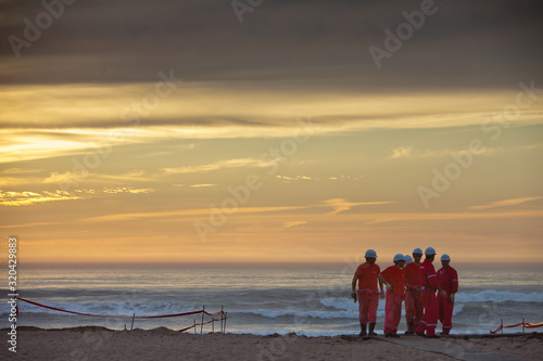 Workers in protective wear discussing at beach against sky during sunset photo