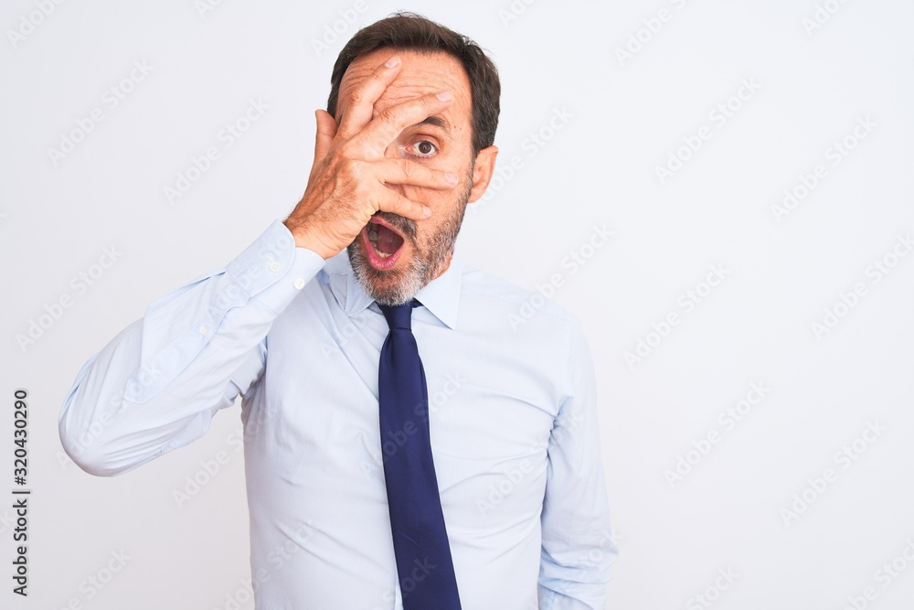 Middle age businessman wearing elegant tie standing over isolated white background peeking in shock covering face and eyes with hand, looking through fingers with embarrassed expression.