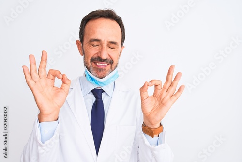 Middle age scientist man wearing coat and medical mask over isolated white background relax and smiling with eyes closed doing meditation gesture with fingers. Yoga concept.