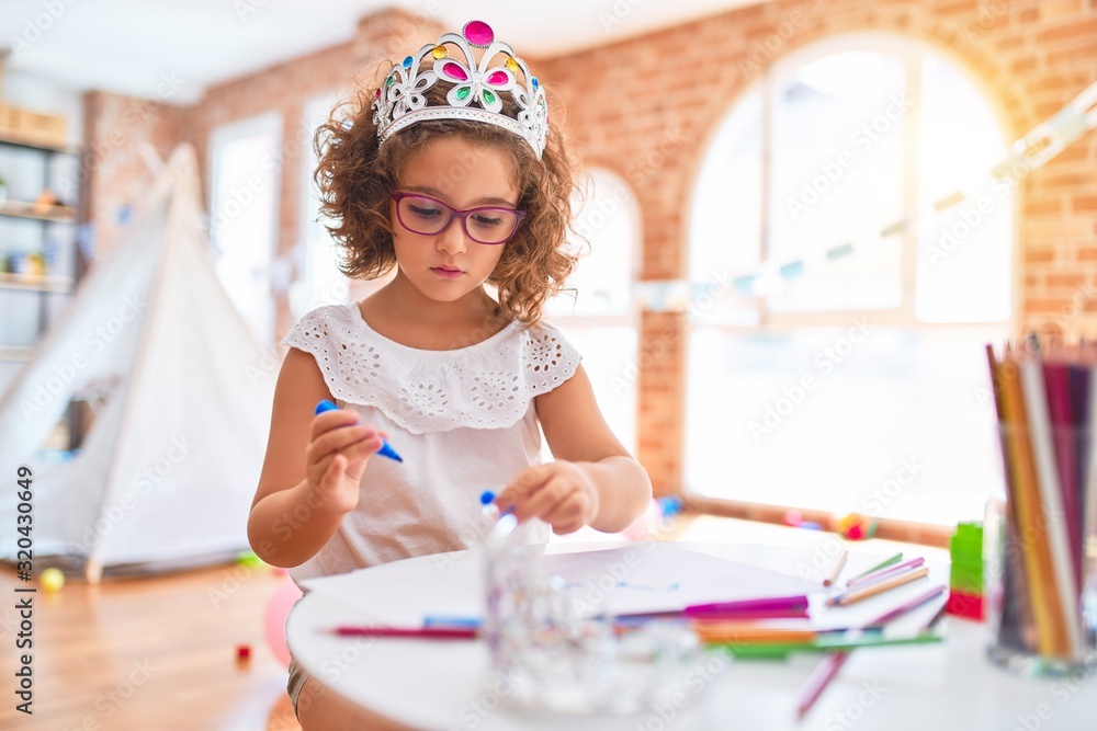 Beautiful toddler wearing glasses and princess crown sitting drawing using paper and marker pen at kindergarten - obrazy, fototapety, plakaty 