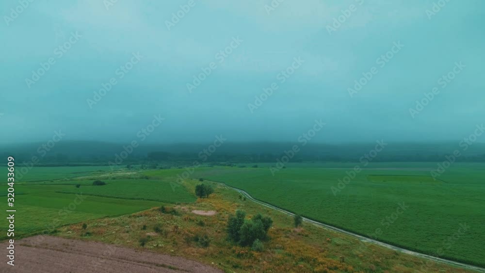 Flight over crop fields and rugged road at the bottom of a mountains in mist at dawn.