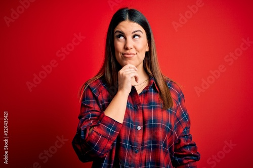 Young beautiful woman wearing casual shirt over red background with hand on chin thinking about question, pensive expression. Smiling with thoughtful face. Doubt concept. © Krakenimages.com