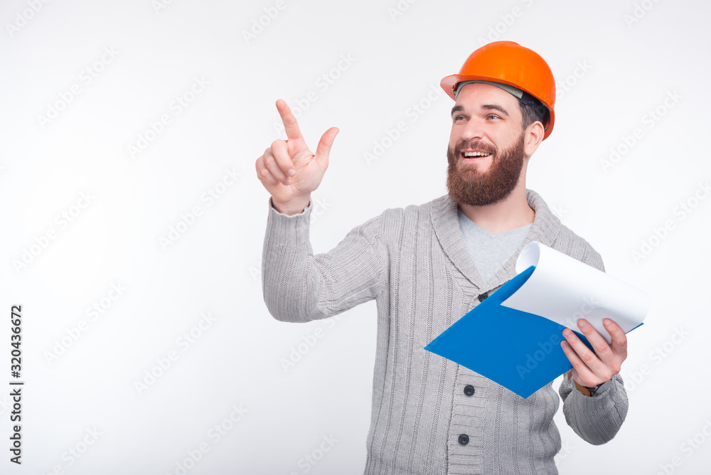 Young bearded architect is pointing or touching the screen while he is holding his board with papers and lists. Young bearded architect is pointing or touching the screen while he is holding his board with papers and lists.