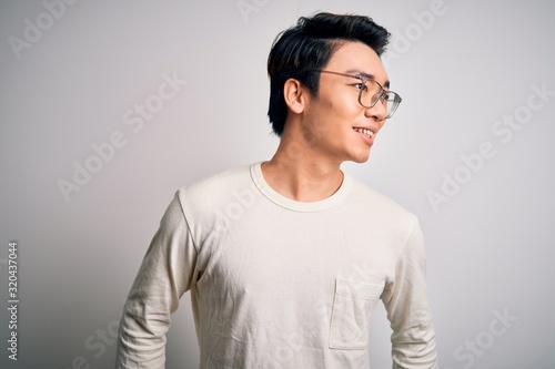 Young handsome chinese man wearing casual t-shirt and glasses over white background looking away to side with smile on face, natural expression. Laughing confident. © Krakenimages.com