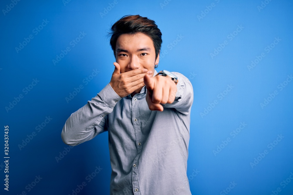 Young handsome chinese man wearing casual shirt standing over isolated blue background laughing at you, pointing finger to the camera with hand over mouth, shame expression