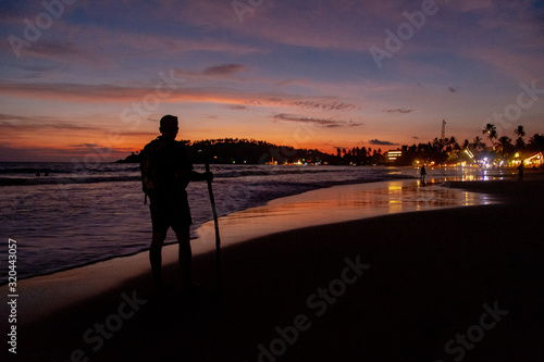 A man walks with a cane along the shore of Mirissa beach at sunset