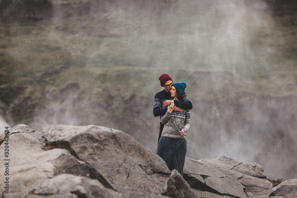 Young stylish couple in love kissing together near famous Icelandic landscape Dettifoss waterfall. Traditional wool sweaters, hat, red hair, gray skirt. Dramatic landscape, cold weather in Iceland. Young stylish couple in love kissing together near famous Icelandic landscape Dettifoss waterfall. Traditional wool sweaters, hat, red hair, gray skirt. Dramatic landscape, cold weather in Iceland.