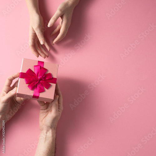 Valentine's Day celebration concept. A nice gift from a loved one. Box with a bow hands of a man and a woman on a delicate pink background. Copy space. Flat lay.Square.
