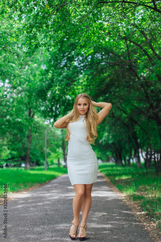 Portrait of a charming blond woman wearing beautiful white dress standing on the road under trees. Portrait of a charming blond woman wearing beautiful white dress standing on the road under trees.