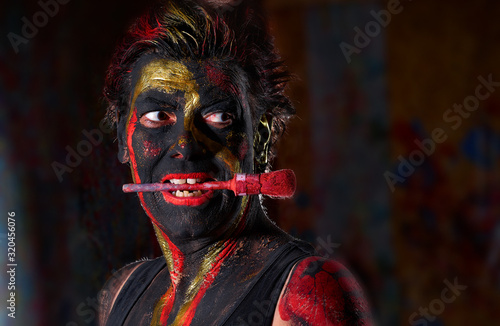 portrait of an attractive European (Caucasian) lady with black, golden and red body painting against dark background - she has a round red brush across her face and looks lasivously to the right side