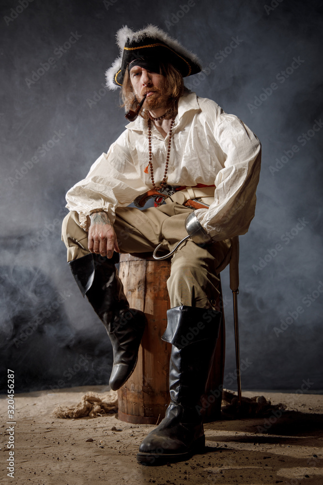 Obraz premium Pirate filibuster sea robber in suit with gun and saber sits on barrel. Concept photo