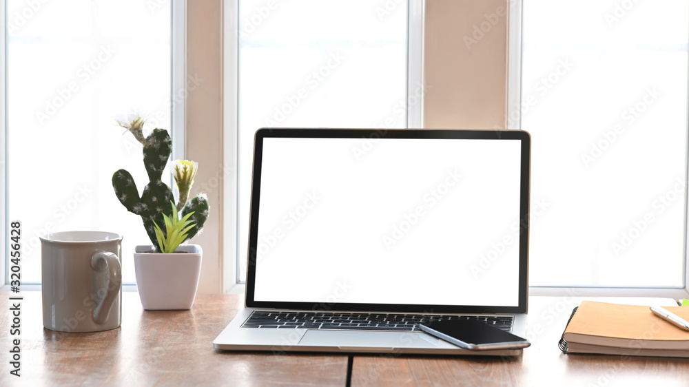 Photo of white blank screen laptop, coffee cup, notebook, pen and potted plant putting together on modern wooden table with light through the window as background. Photo of white blank screen laptop, coffee cup, notebook, pen and potted plant putting together on modern wooden table with light through the window as background.