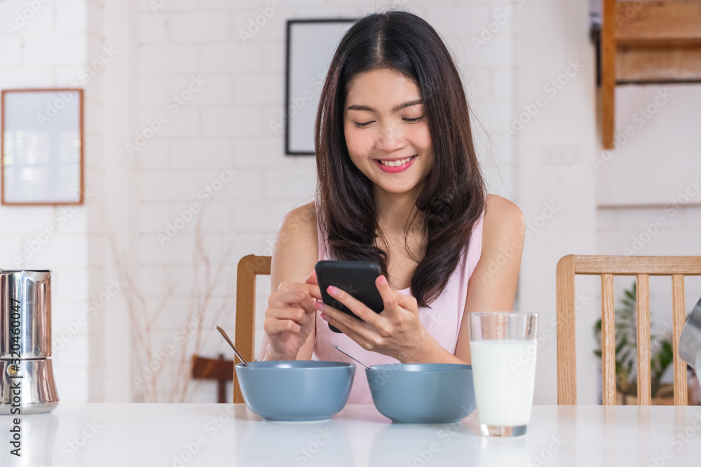 Asian woman using mobile and eating cereal breakfast on kitchen table in morning.daily lifestyle Asian woman using mobile and eating cereal breakfast on kitchen table in morning.daily lifestyle