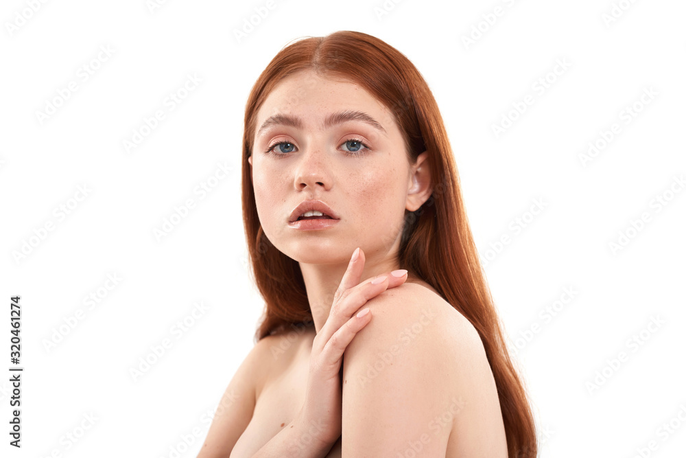 Sensual beauty. Portrait of young and cute half naked red-haired woman with freckles on face looking at camera and keeping hand on her shoulder while standing against white background Sensual beauty. Portrait of young and cute half naked red-haired woman with freckles on face looking at camera and keeping hand on her shoulder while standing against white background