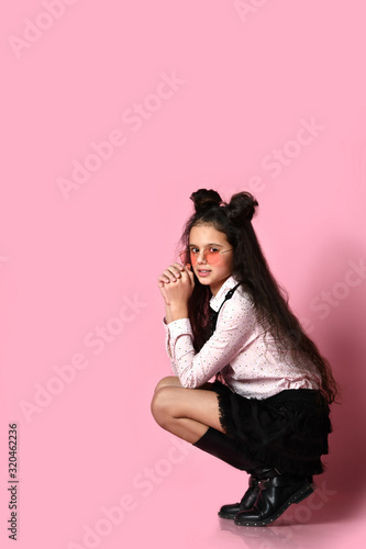 Teenage kid with long hair, in sunglasses, white blouse, black skirt and boots. She posing squatting on pink background. Close up