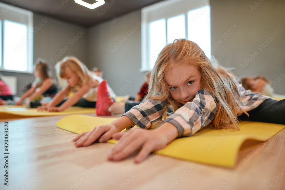 Close up portrait of a little cute girl sitting on yoga mat and stretching her body. Group of children doing gymnastic exercises in the dance studio