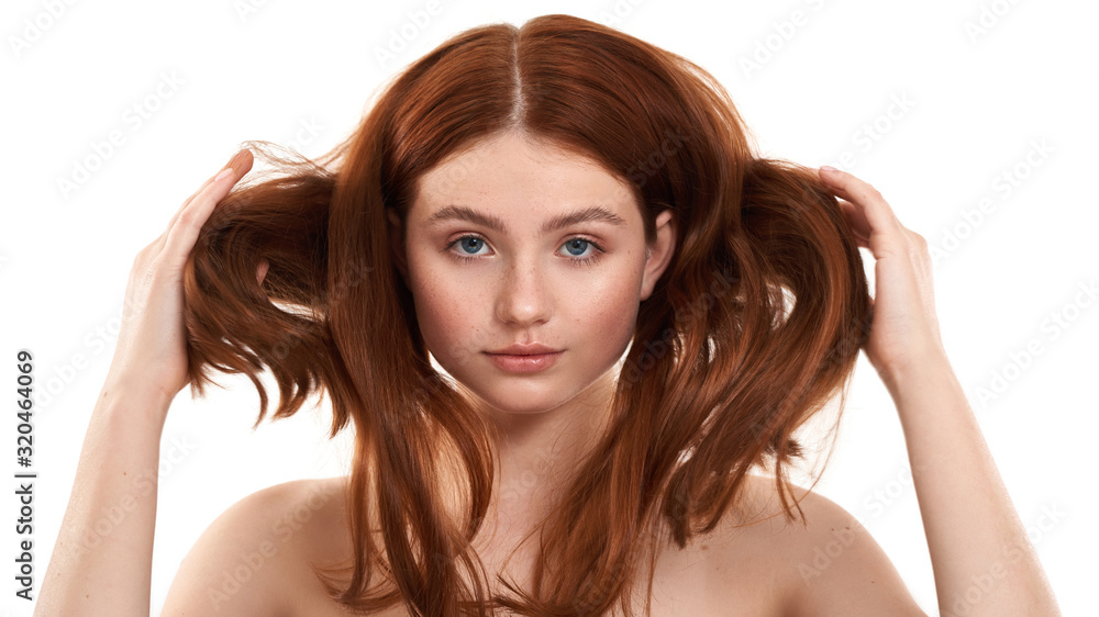 Fototapeta premium Power of hair. Portrait of a young gorgeous redhead woman playing with her long and healthy ginger hair while standing against white background. Natural beauty