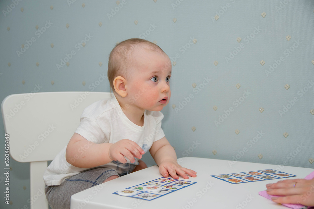 A small child sits at a white table and plays in a children's lotto. The kid studies animals on cards, plays a board game. A small child sits at a white table and plays in a children's lotto. The kid studies animals on cards, plays a board game.