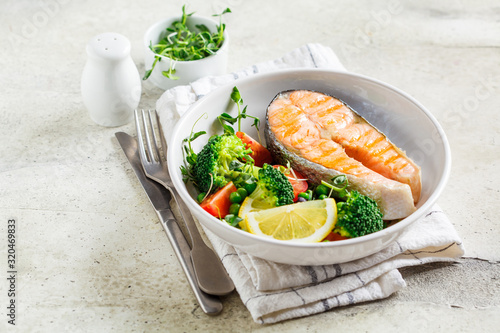 Grilled salmon steak with broccoli and tomatoes in white plate on gray background, copy space. Diet food concept.