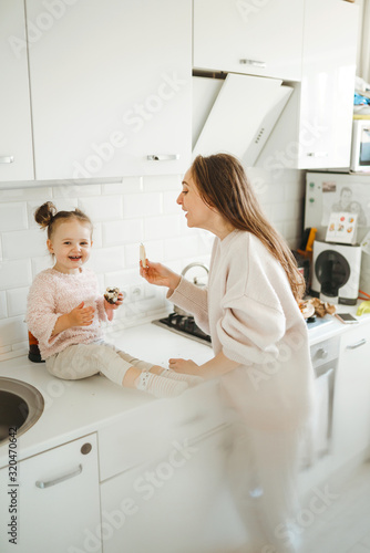 Mother and little daughter have fun together in the sunny kitchen. Little girl sitting on table top and eats a bakery products with mom. Happy loving family.