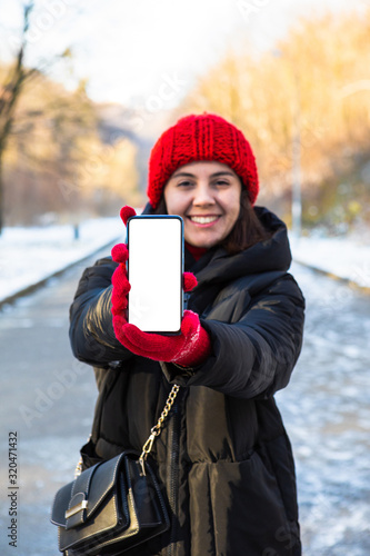 young smiling woman in winter outfit holding phone with white blank empty screen