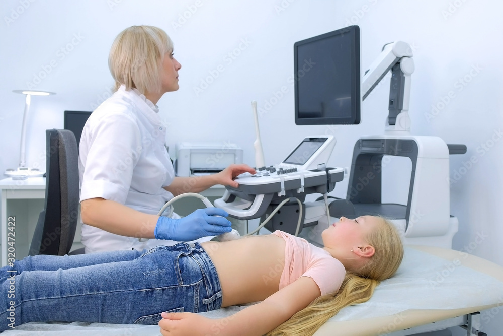 Doctor is making abdominal ultrasound for child girl using scanner machine in clinic, side view. Woman runs ultrasound sensor over patient tummy in clinic. Diagnostic examination of internal organs.