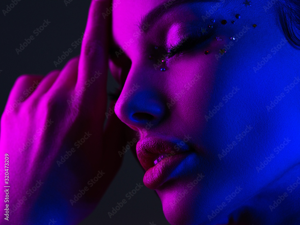 Portrait of the girl is highlighted in bright blue and purple light. Face of a beautiful model. Colorful portrait of a girl on a dark background. Shiny sequins on the face. Art portrait. Sexy woman. Portrait of the girl is highlighted in bright blue and purple light. Face of a beautiful model. Colorful portrait of a girl on a dark background. Shiny sequins on the face. Art portrait. Sexy woman.