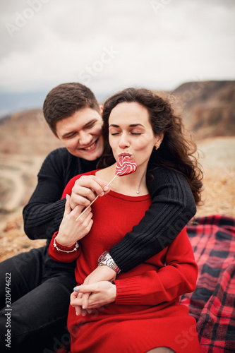 loving couple a boy and a girl sitting on a red-black plaid, gently hugging in their hands caramel on a stick on a background of a lake in the shape of a heart in softfocus
