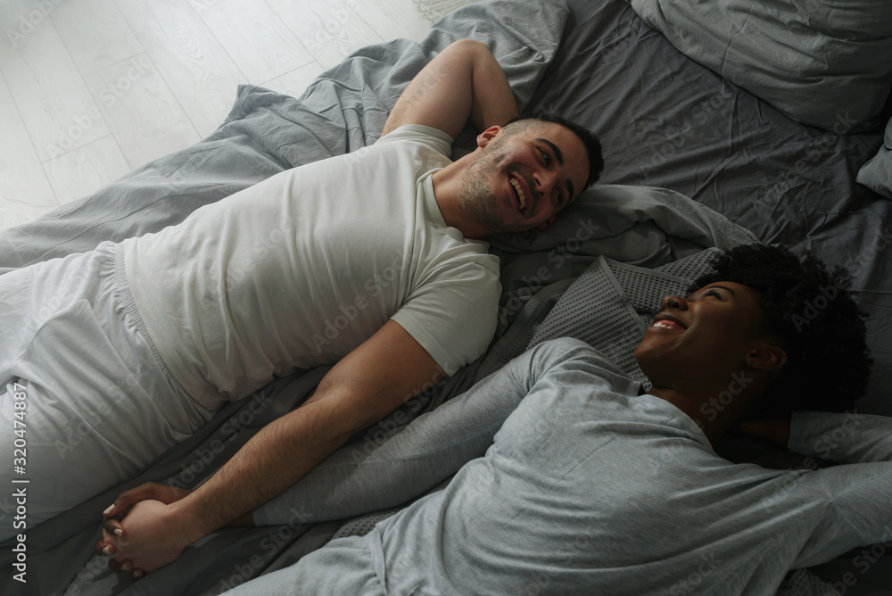 Young smiling mixed race couple lying together in bed in the morning. Love couple holding hands in the bed. Young smiling mixed race couple lying together in bed in the morning. Love couple holding hands in the bed.