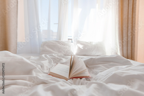 Book on bed with white linen in front of window with morning light through curtains