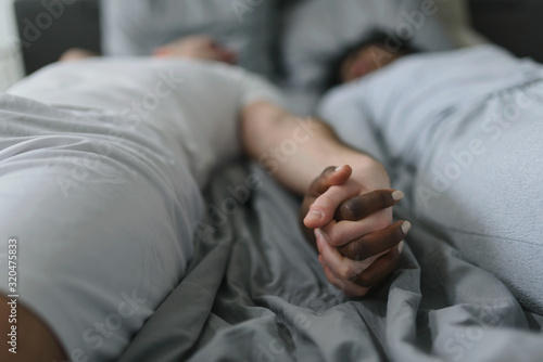 Young smiling mixed race couple lying together in bed in the morning. Love couple holding hands in the bed. © lordn