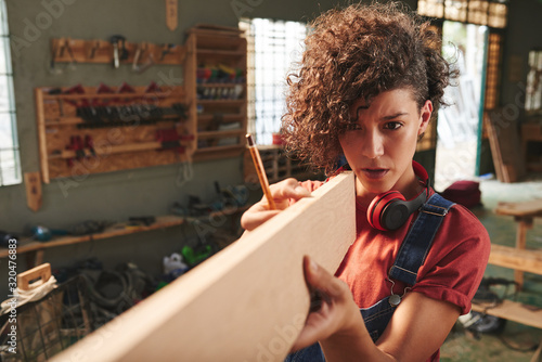 Young concentrated female carpenter with curly hair holding wooden plank and estimating its length before sawing photo